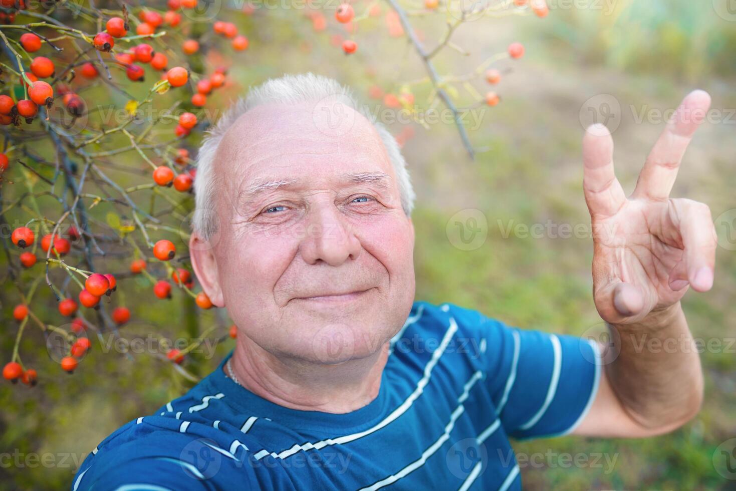 positive retired grandfather, makes selfie photo with a smartphone in the park. an elderly man walks in nature.
