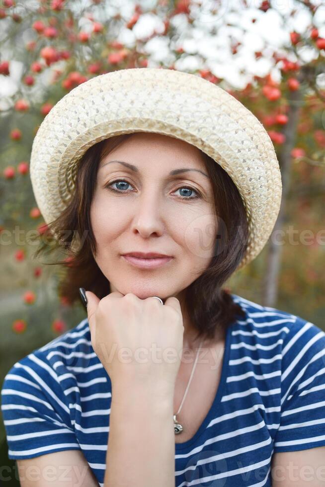 Image of beautiful middle-aged woman wearing straw hat laughing and looking at camera while walking in autumn park photo