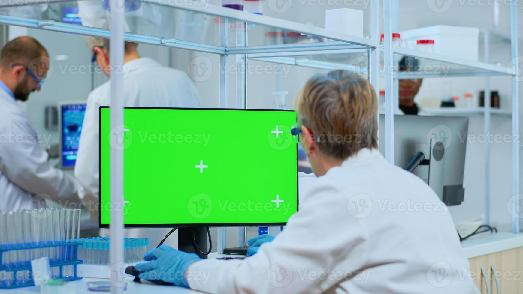 Elderly researcher looking at chroma key display in modern equipped lab. Multiethnic team of microbiologists doing vaccine research writing on device with green screen, isolated, mockup display. photo