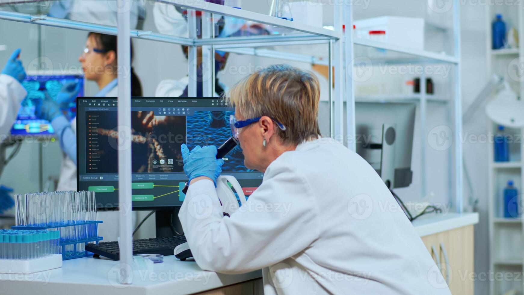 Senior scientist looking through a microscope in modern equipped lab. Multiethnic team examining virus evolution using high tech for scientific research of vaccine development against covid19. photo