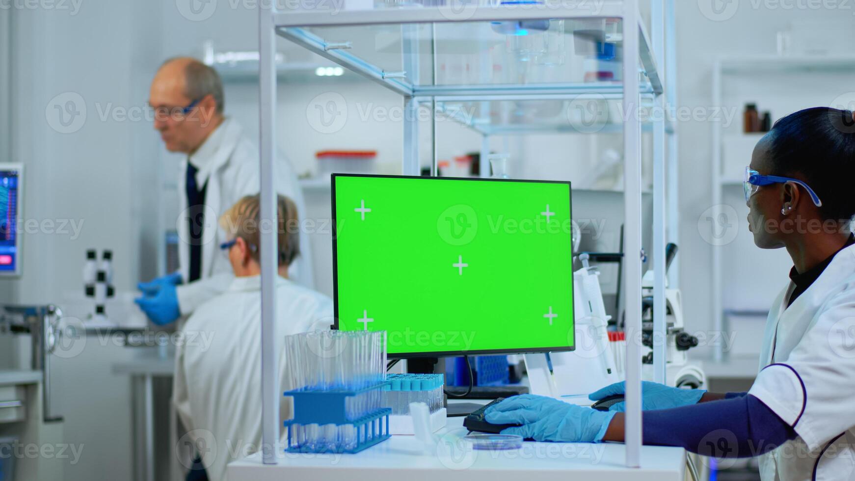 Black woman researcher looking at chroma key computer in modern equipped lab. Multiethnic team of microbiologists doing vaccine research writing on device with green screen, isolated, mockup display. photo
