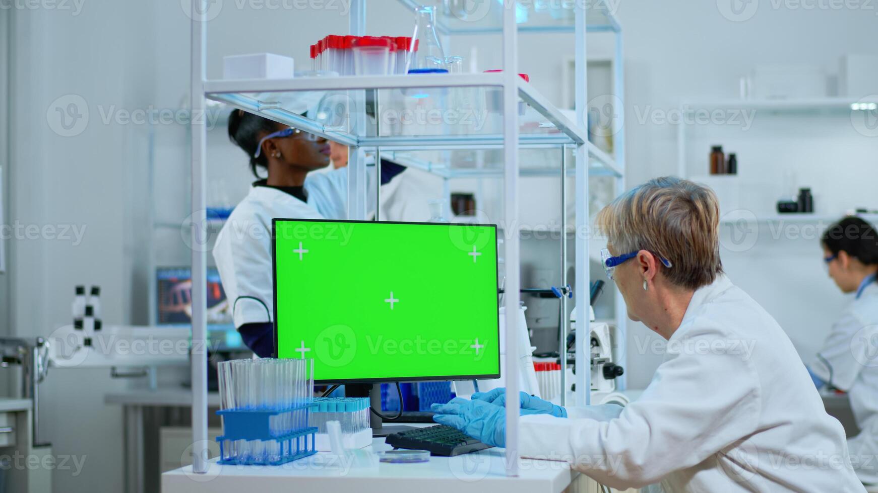 Senior doctor working at computer with green screen in modern equipped lab. Multiethnic team of microbiologists doing vaccine research writing on device with chroma key, isolated, mockup display. photo