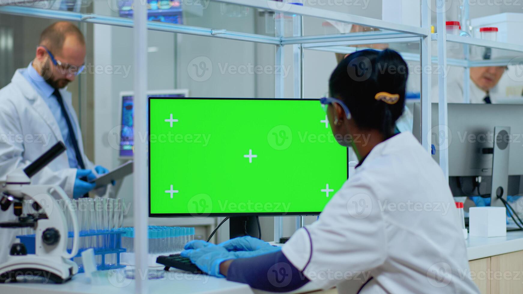African doctor working at computer with green screen in modern equipped lab. Multiethnic team of microbiologists doing vaccine research writing on device with chroma key, isolated, mockup display. photo
