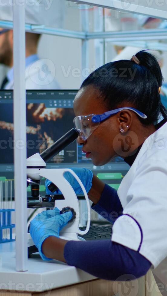 Black woman chemist analysing reaction of virus on microscope in laboratory. Multiethnic team examining vaccine evolution using high tech for scientific research, treatment development against covid19 photo