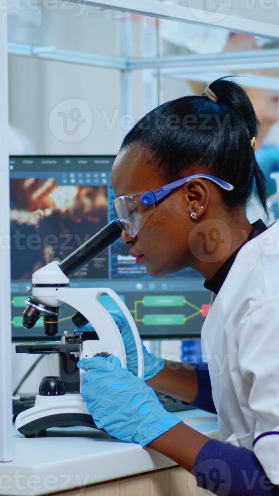 Black biochemist woman sitting in modern equipped lab analysing blood tests. Multiethnic team examining virus evolution using high tech for scientific research of treatment development against covid19 photo