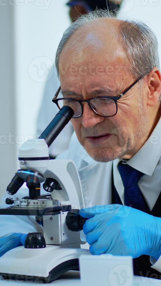 Laboratory scientist conducting experiment looking at microscope in busy modern lab. Eldery man working with various bacteria tissue, blood samples, concept of pharmaceutical research for antibiotics photo