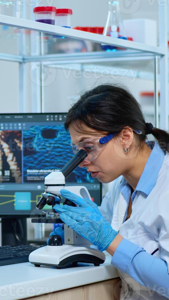 Scientist team checking virus development using microscope and writing analysis results on tablet in modern equipped lab. Multiethnic group examining vaccine evolution using high tech for research. photo