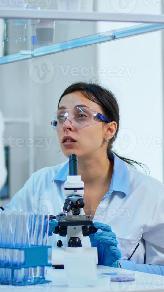 Team medical scientist conducting DNA experiments under digital microscope writing results into tablet working in science laboratory. Caucasian lab engineer in white coat analysing vaccine development photo