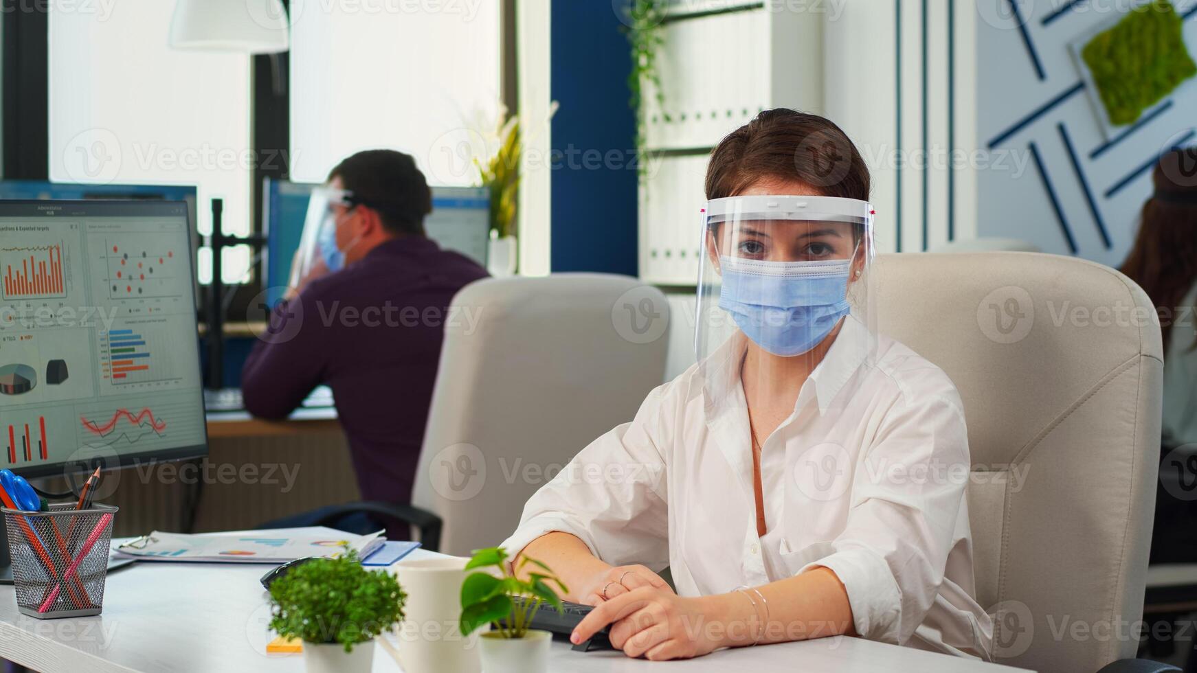 Businesswoman with visor and mask talking in front of camera having call with partners. Pov of manager speaking with remotely team during online conference while colleagues working in background photo