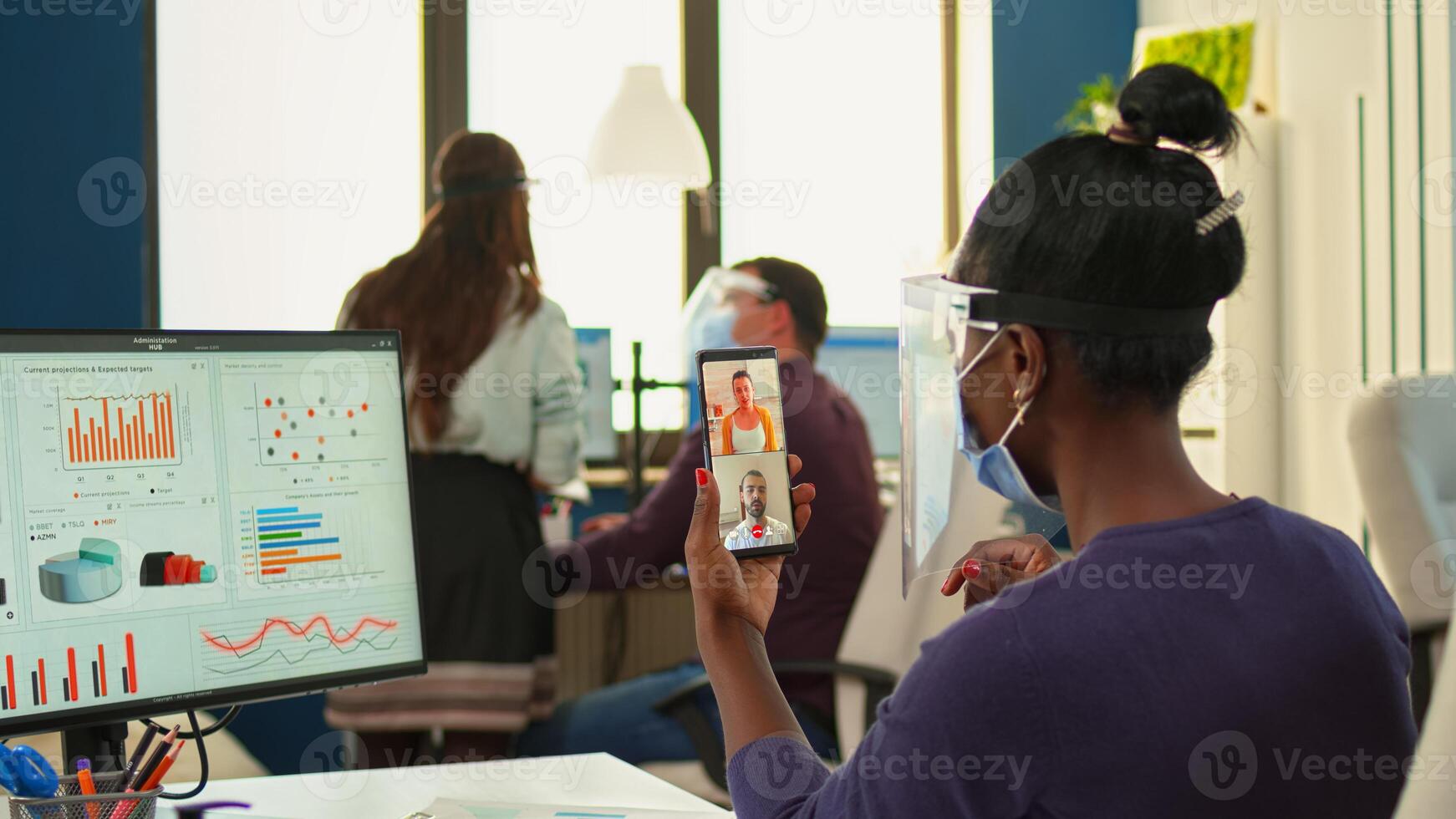 Black business woman with face protection against covid-19 pandemic having online meeting on smartphone sitting in modern workplace. Multiethnic coworkers working respecting social distance in business photo