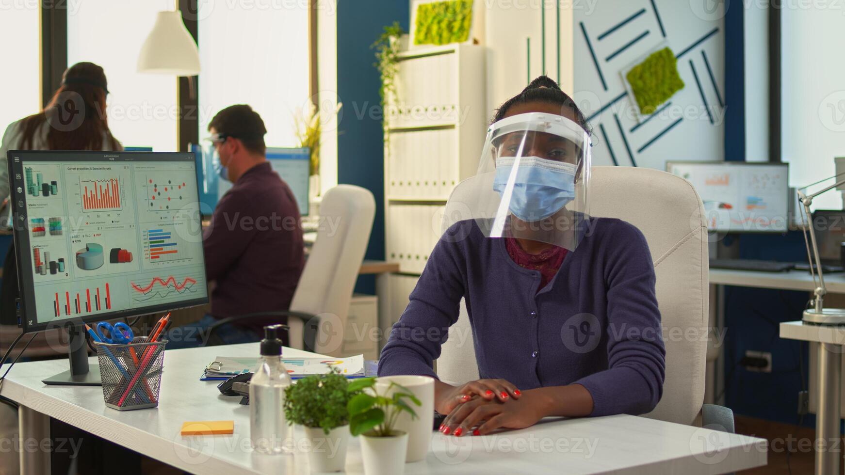African businesswoman with visor and mask talking at camera having meeting with remote partners. Pov of woman speaking with team during online conference while colleagues working in background photo