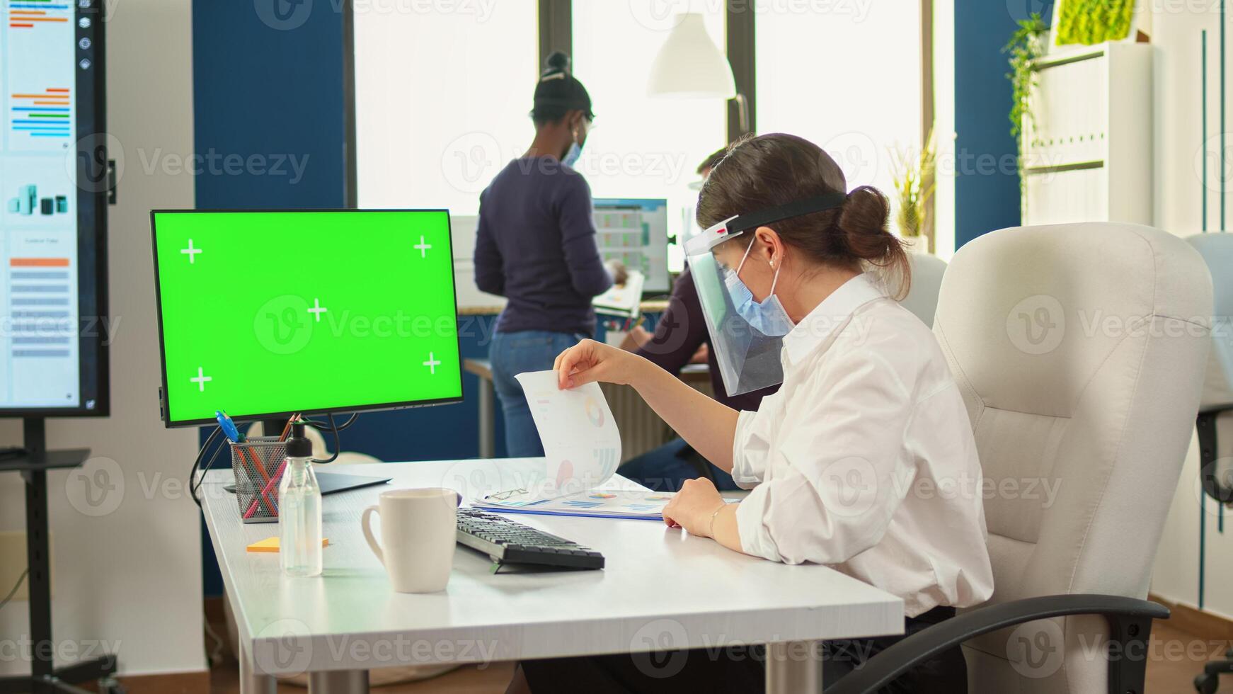 Company manager with face mask typing on computer with green screen in new normal business office respecting social distance during global pandemic. Woman Looking at mockup, copy space, chroma display photo