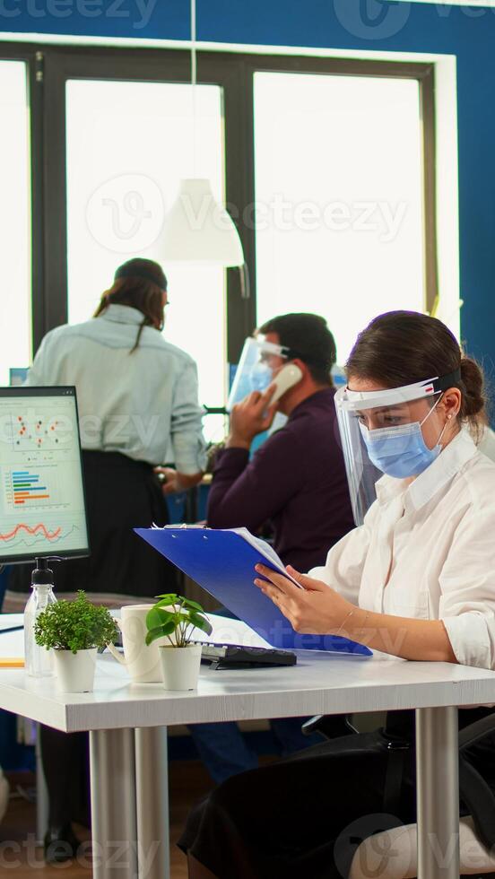 Teamworkers in new normal office making financial strategy wearing protective mask. Employees with visors working in corporate company workspace respecting social distance analysing data and graphs. photo