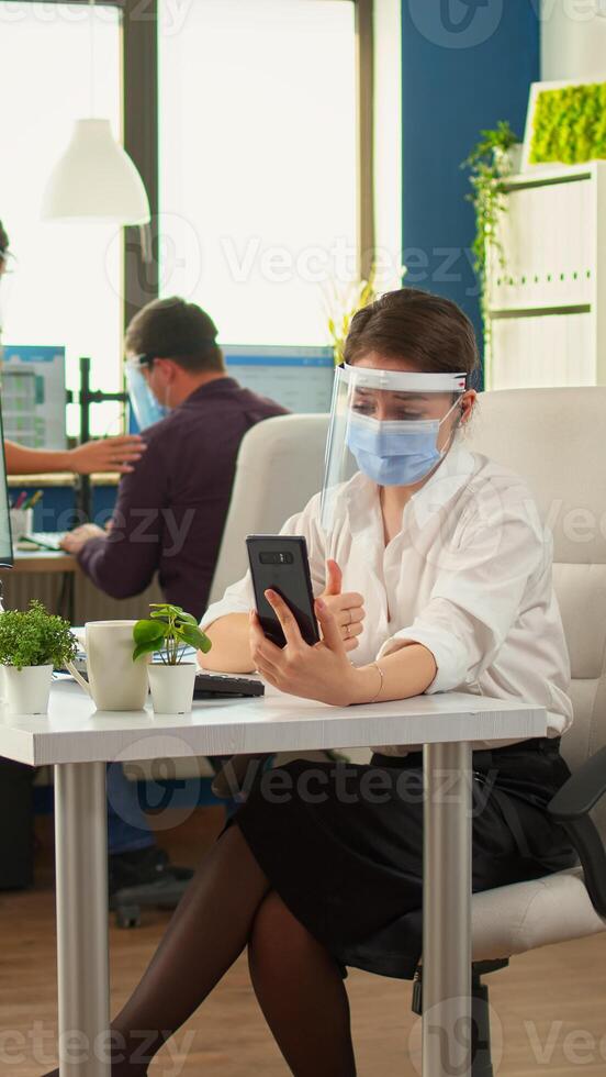 Entrepreneur wearing visor against coronavirus using phone with wireless headphone for videocall talking with remotely team sitting in new normal office. Colleagues working respecting social distance photo