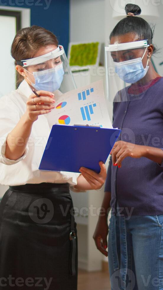 Company manager with visor against coronavirus explaining to african colleague project plan standing in front of desk holding clipboard. Multiethnic business team working respecting social distance. photo