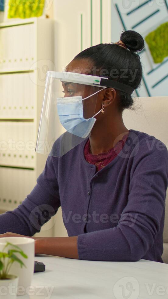 African company manager with face mask writing on computer and looking on camera in new normal business financial office respecting social distance. Multiethnic colleagues working in background. photo