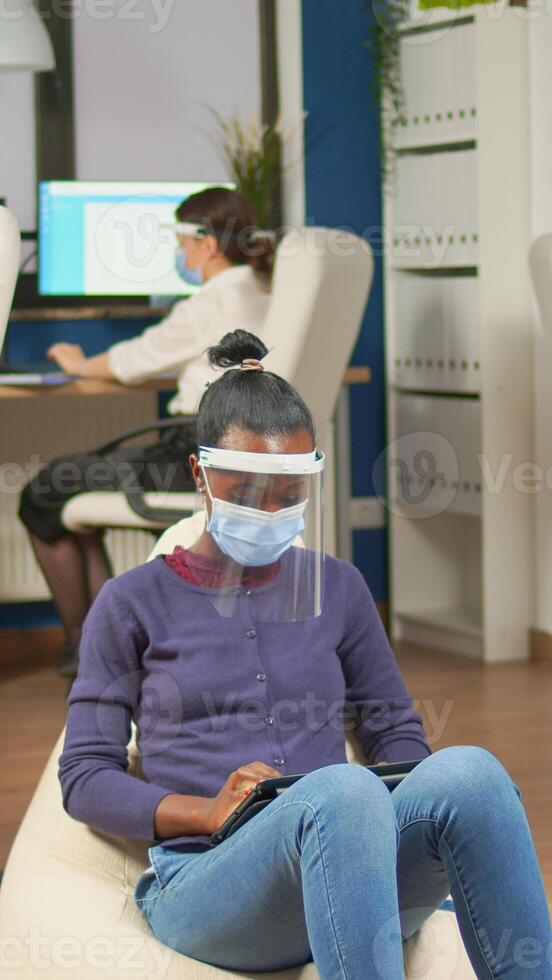 African employee with protection mask against covid-19 pandemic sitting on armchair in middle of office room holding digital tablet. Multiethnic business team working respecting social distance photo