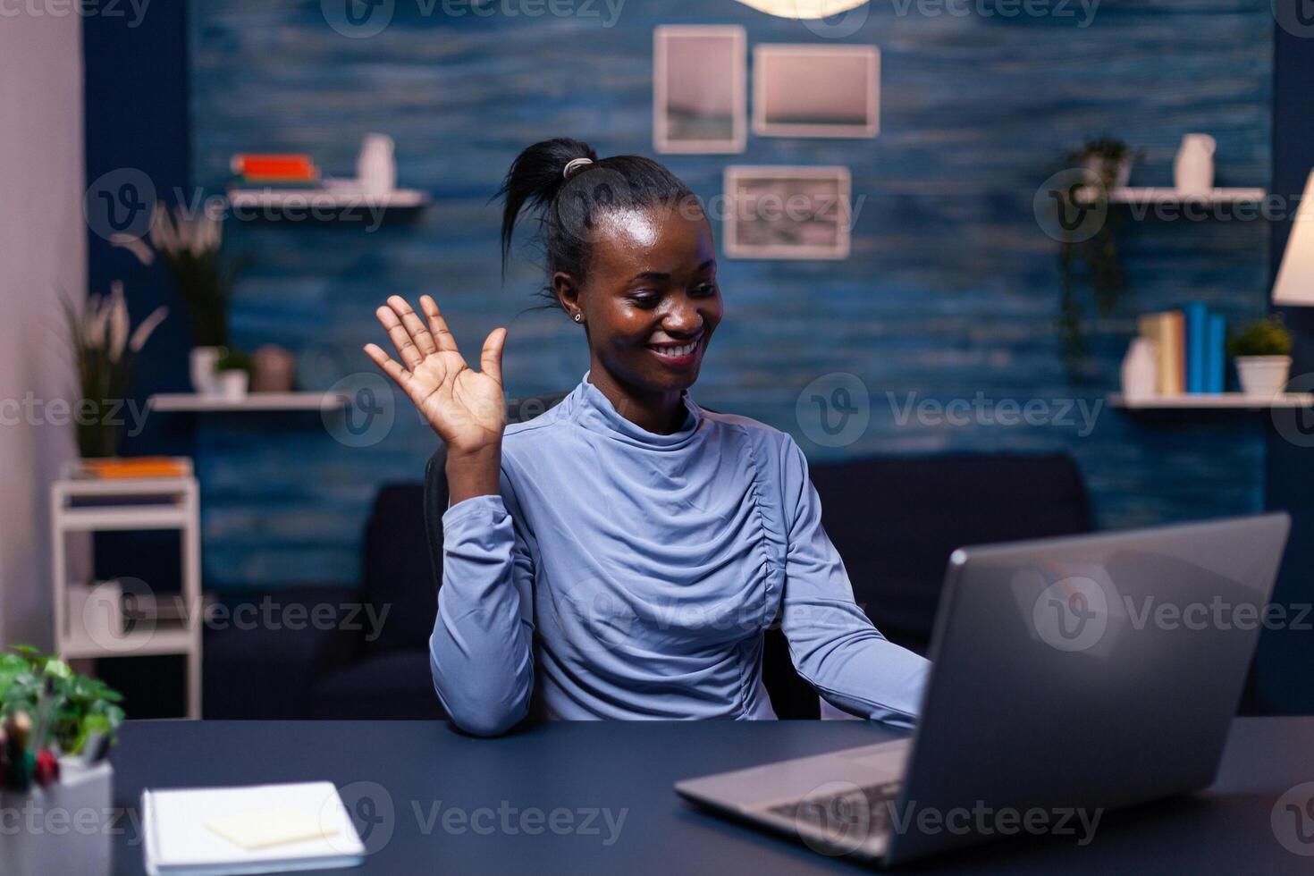 African business woman waving at clients in the course of virtual meeting sitting at desk in home office late at night. Black freelancer working with remotely team chatting virtual online conference. photo