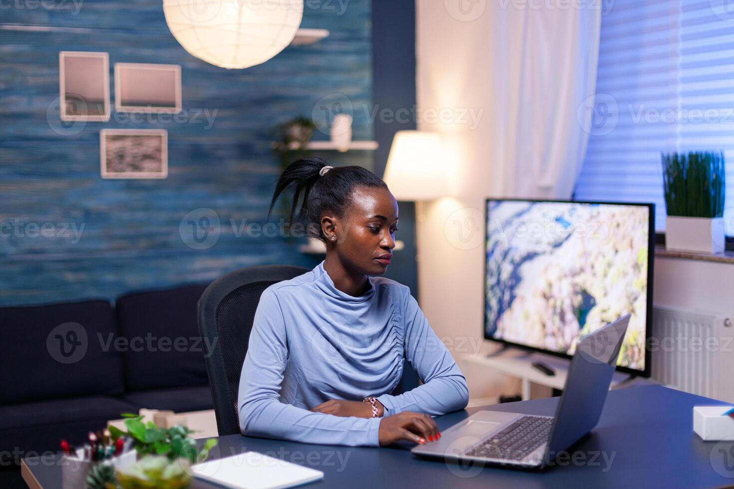 Dark skinned freelancer reading documentation on laptop computer in living room late at night. Black entrepreneur sitting in personal workplace writing on keyboard. photo