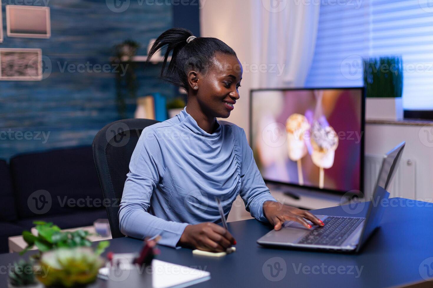 Dark skinned woman taking notes while working on deadline using laptop sitting at desk from home in the evening. Black entrepreneur sitting in personal workplace writing on keyboard. photo