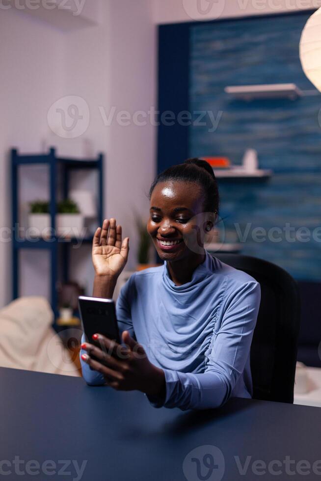 Dark skinnned woman saying hello in the course of conference sitting at desk late at night in office. Black freelancer working with remotely team chatting virtual online conference. photo