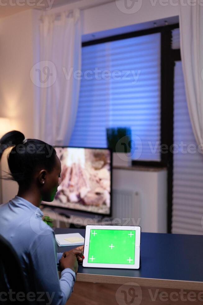 African entrepreneur sitting at desk in home office holding tablet pc with green screen working on deadline. Using mockup chroma key display computer. photo