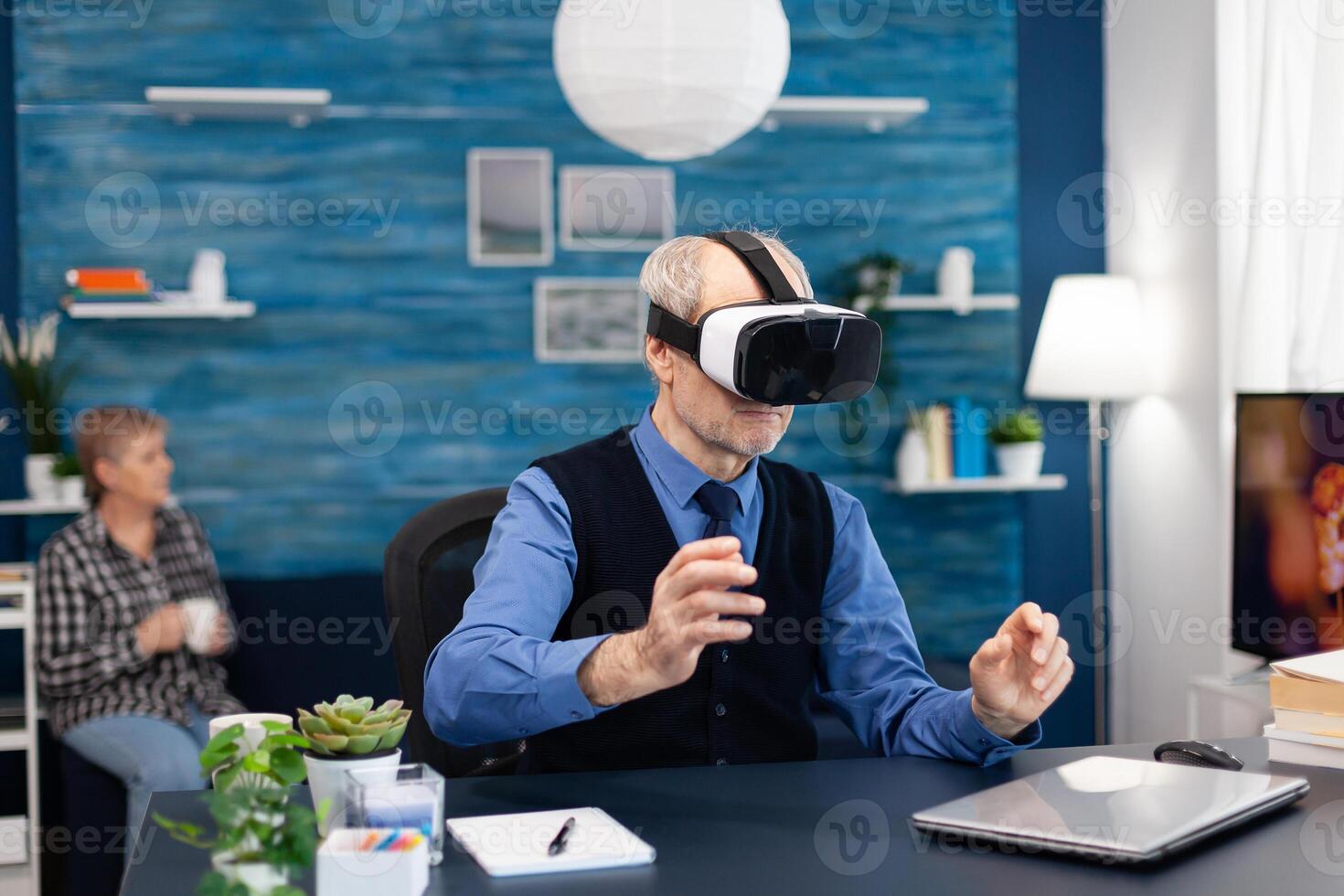Senior man wearing virtual reality headset doing hand gesturing. Elderly man pensioner in living room sitting at desk with futuristic goggles for augmented cyberspace wife watching tv. photo