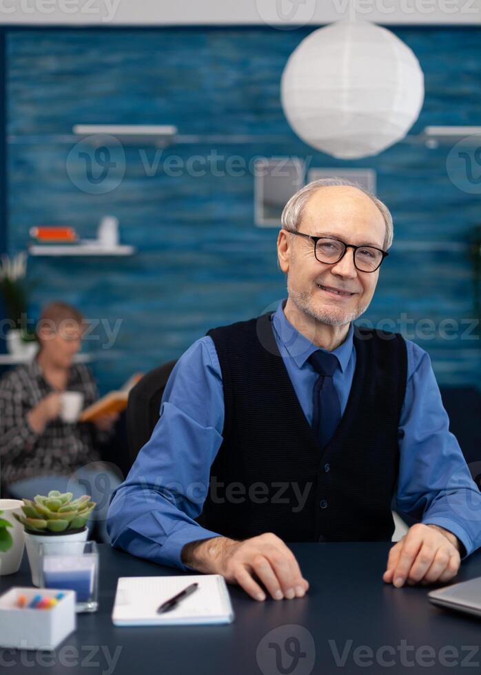 Cheerful senior businessman with gray hair. smiling at camera Elderly man entrepreneur in home workplace using portable computer sitting at desk while wife is reading a book sitting on sofa. photo