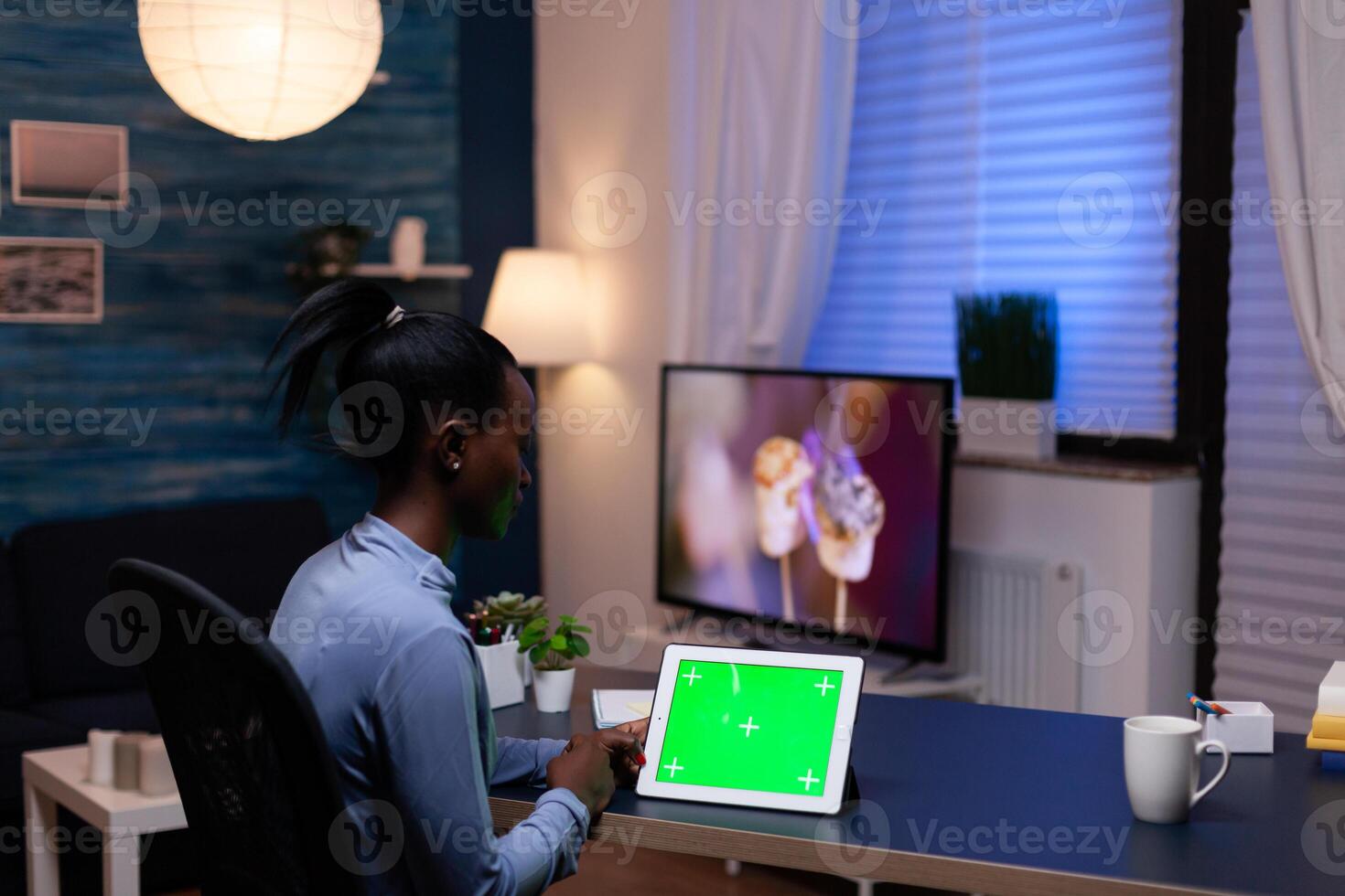 Black entrepreneur woman working on project deadline from home at night in home office holding tablet pc with green screen. Using mockup chroma key display computer. photo