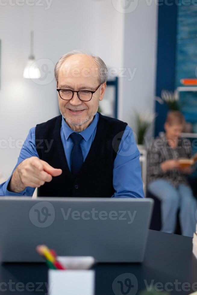 Happy senior man pointing at laptop during conference. Cheerful pensioner with gray hair smiling at laptop webcam while chatting sitting at desk . photo