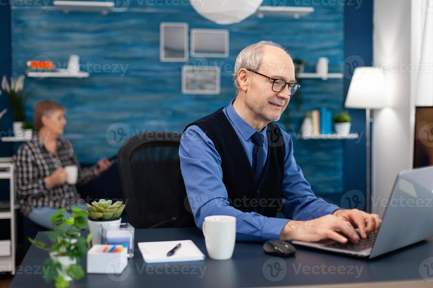 Portrait of senior man typing on laptop keyboard working on project. Elderly man entrepreneur in home workplace using portable computer sitting at desk while wife is holding tv remote. photo