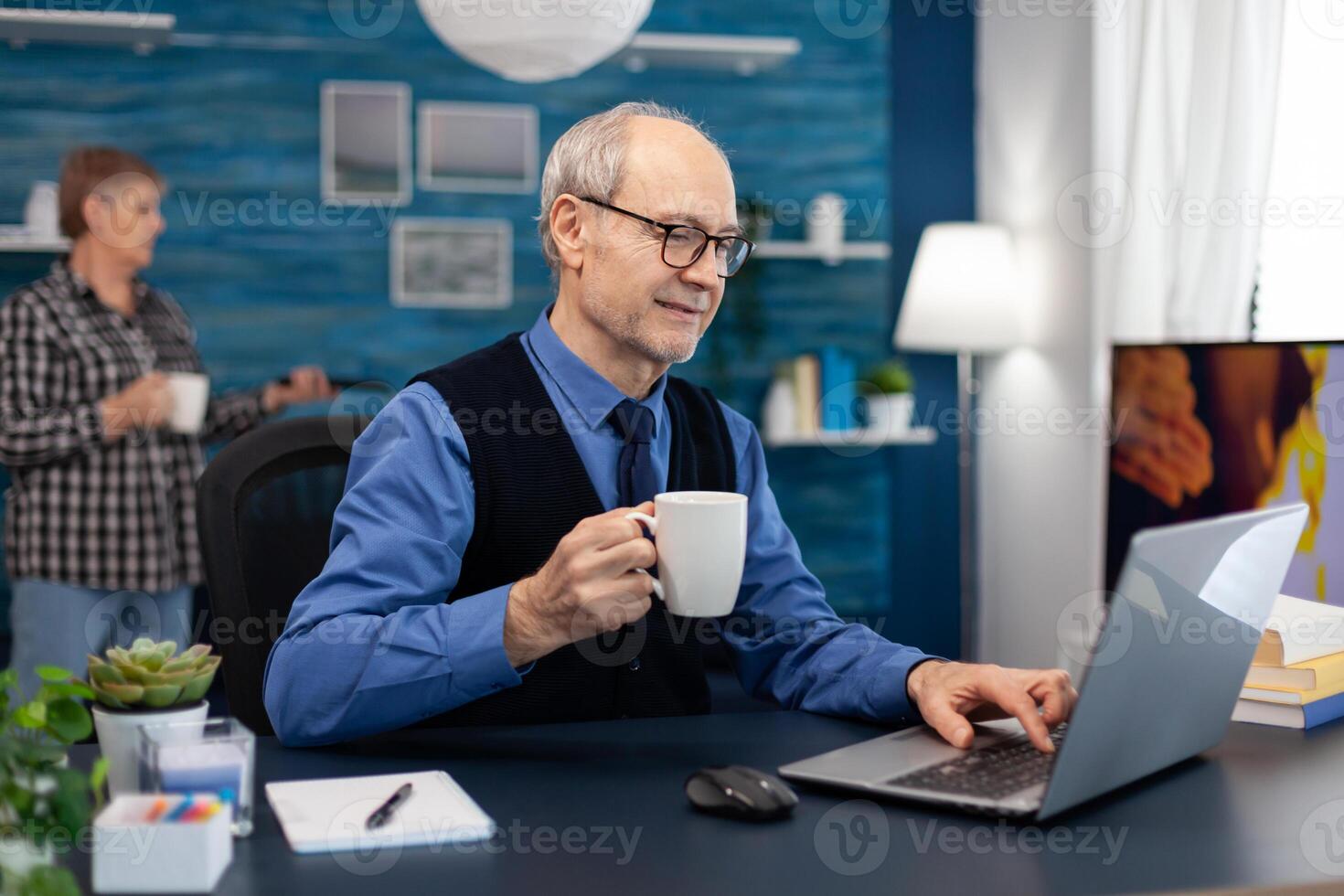 Senior businessman holding cup of coffee working on laptop. Elderly man entrepreneur in home workplace using portable computer sitting at desk while wife is holding tv remote. photo