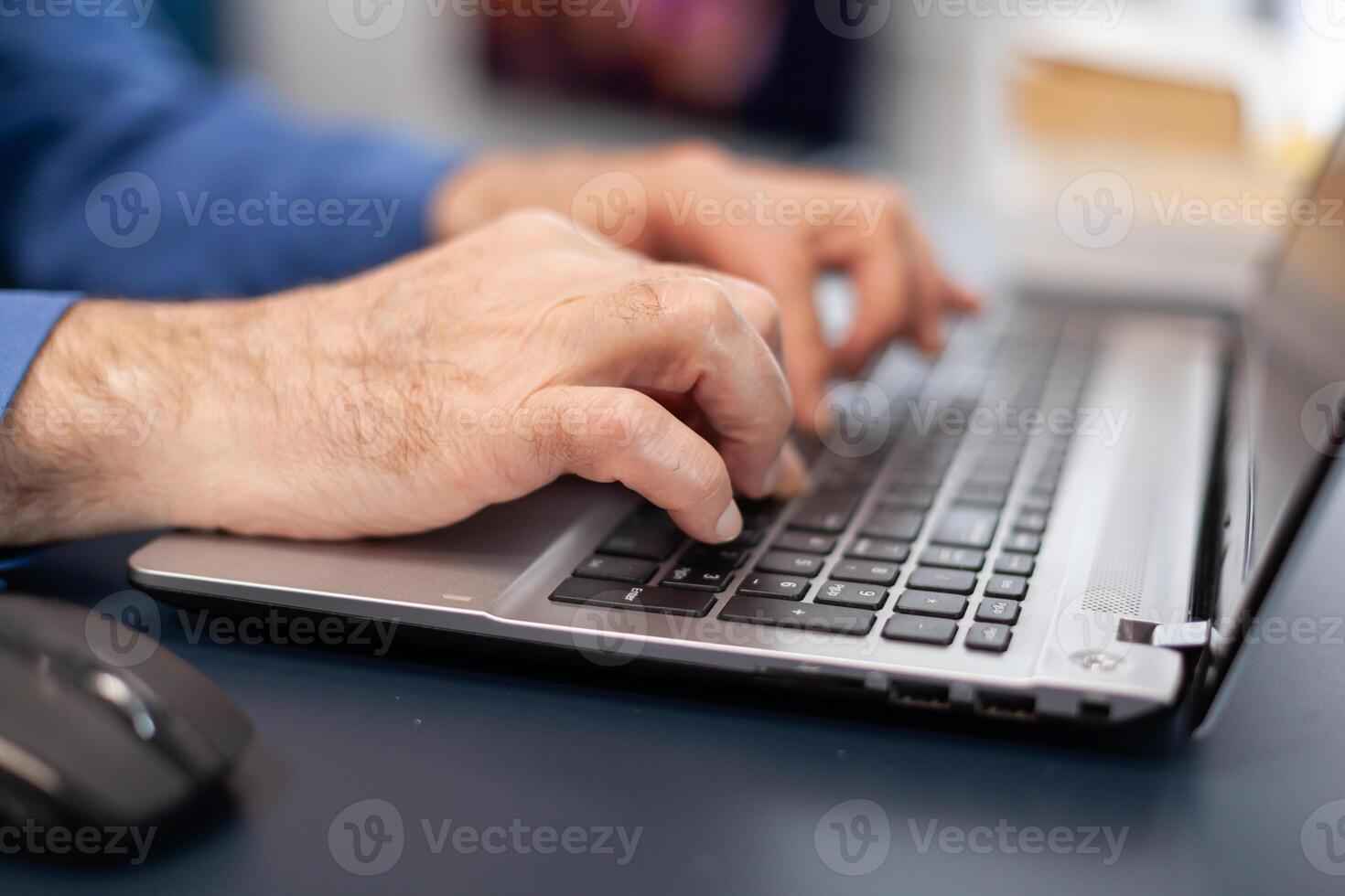 Close up of senior man hands typing on laptop keyboard. Elderly man entrepreneur in home workplace using portable computer sitting at desk while wife is holding tv remote photo
