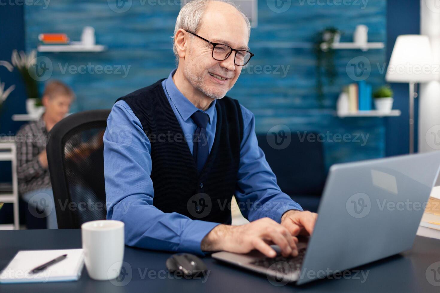 Smart senior businessman working on laptop wearing a tie and glasses. Elderly man entrepreneur in home workplace using portable computer sitting at desk while wife is holding tv remote. photo