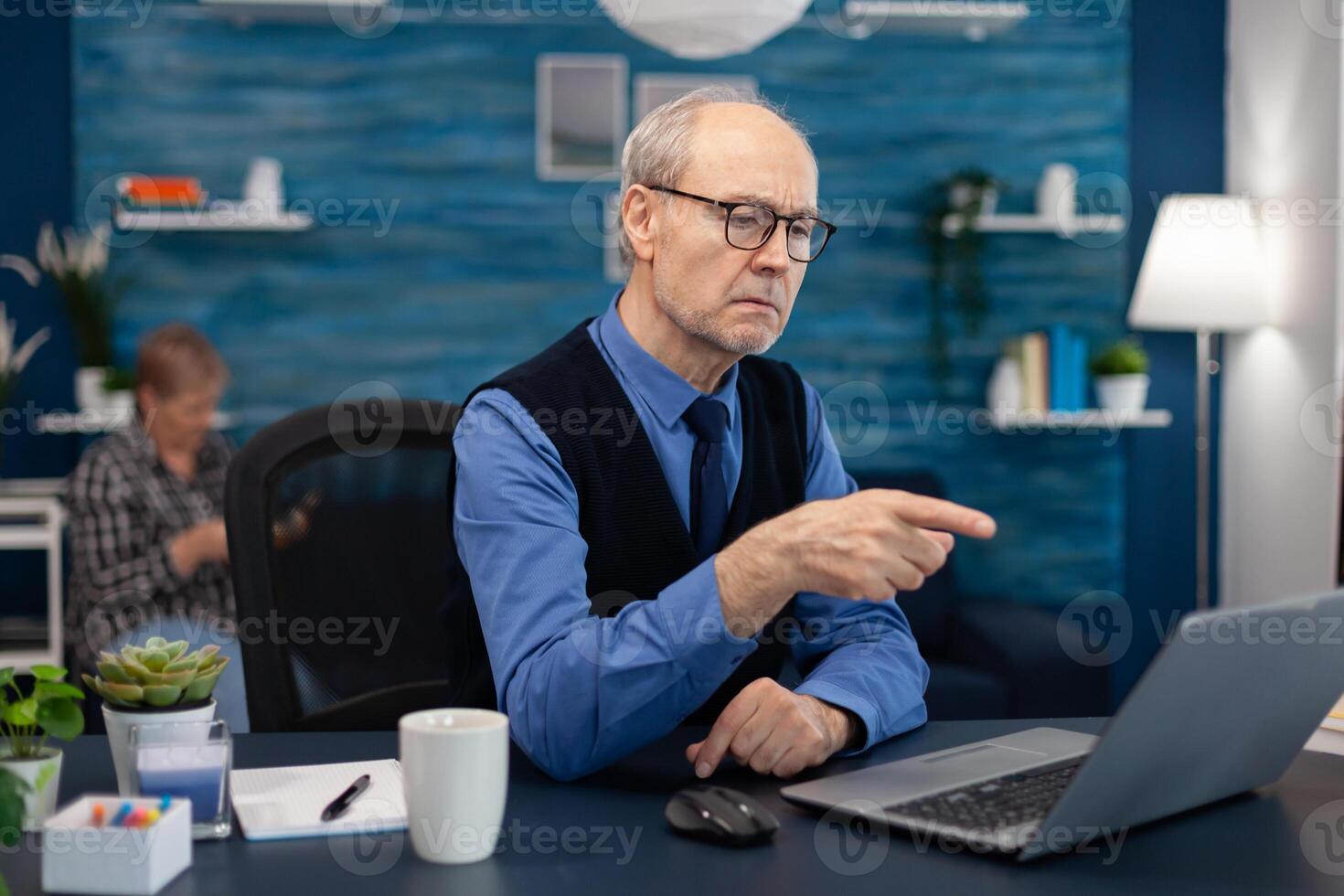 Thoughtfull senior businessman pointing at laptop whille working from home. Elderly man entrepreneur in home workplace using portable computer sitting at desk while wife is holding tv remote. photo