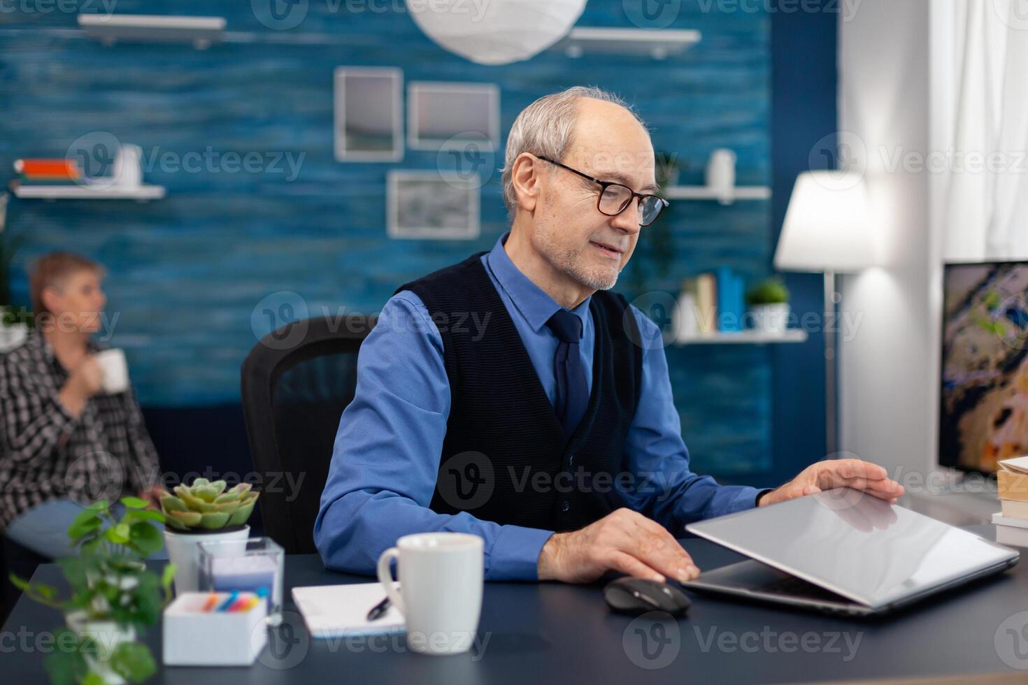 Businessman with gray hair opening laptop to start working. Elderly man entrepreneur in home workplace using portable computer sitting at desk while wife is holding tv remote. photo