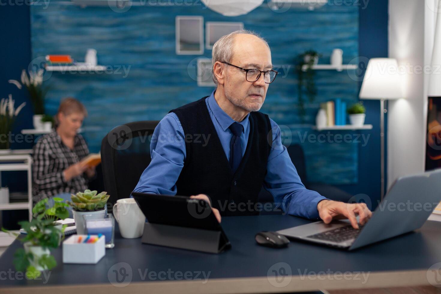 Senior manager working on presentation using laptop and tablet pc sitting at office. Elderly man entrepreneur in home workplace using portable computer sitting at desk while wife is reading a book sitting on sofa. photo