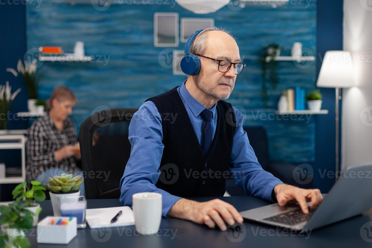 Senior businessman listening music wearing headphones working on laptop. Elderly man entrepreneur in home workplace using portable computer sitting at desk while wife is reading a book sitting on sofa. photo