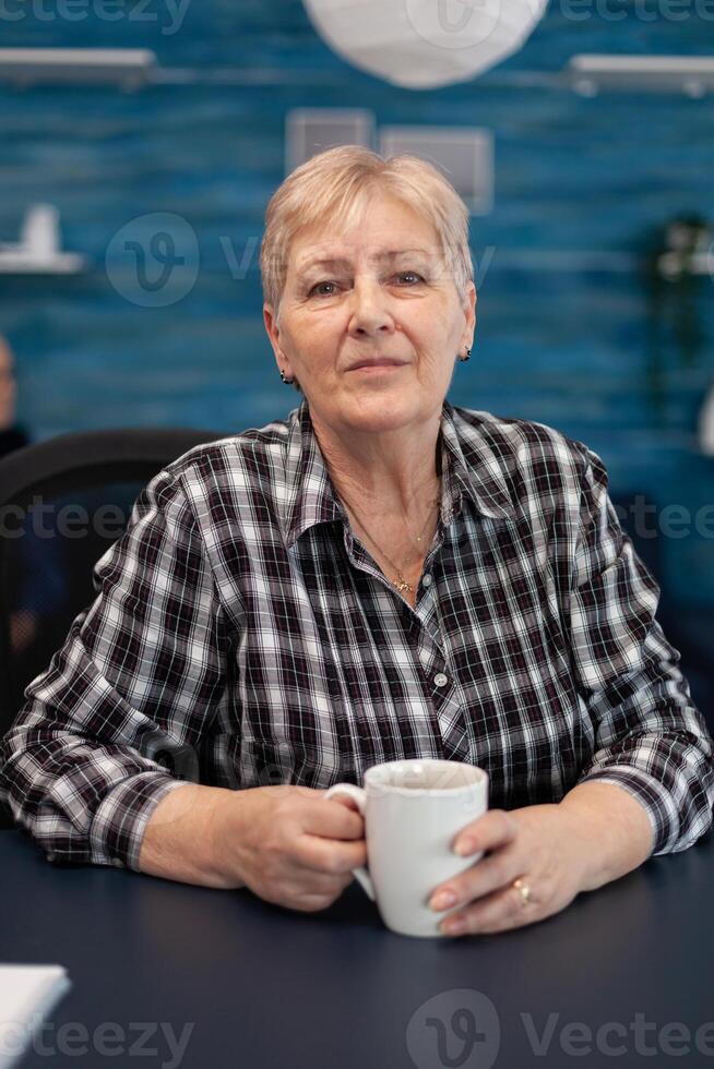 Portrait of senior woman looking at camera while enjoying a mug of coffee. Healthy happy elderly woman in home living room enjoying her retirement photo