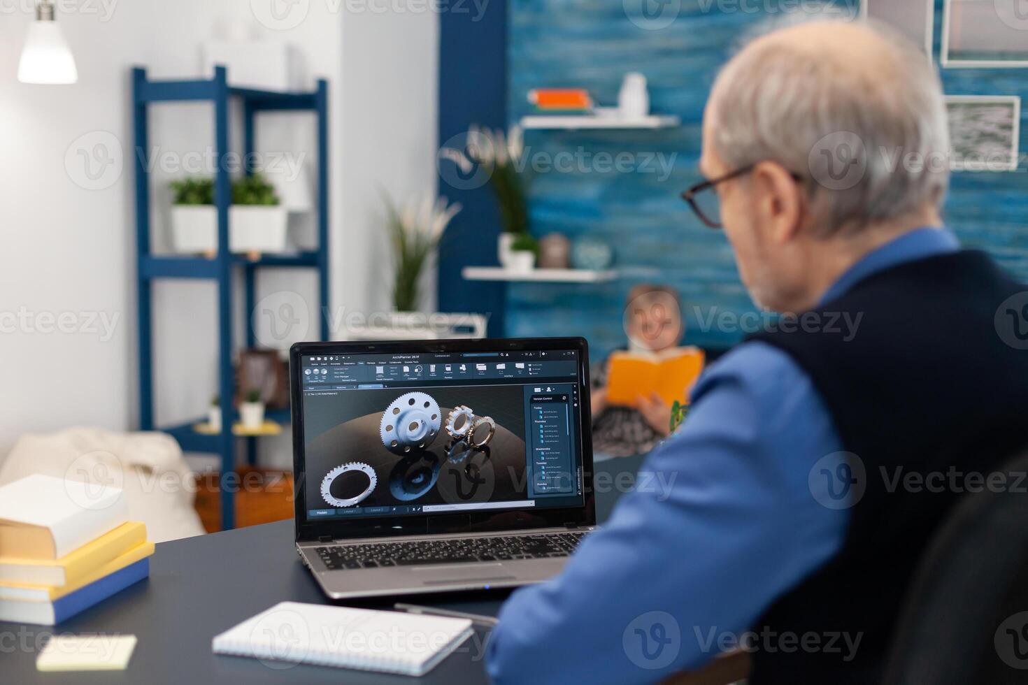 Senior engineer working on deadline sitting at desk. Experienced engineer working from home office on mechanical project using laptop computer technology and wife is reading a book on sofa. photo