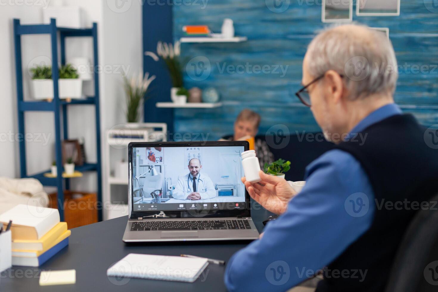 Senior man showing doctor pills bottle at webcam during call. Elderly man discussing with healthcare practitioner in the course of remote call and wife is reading a book on sofa. photo