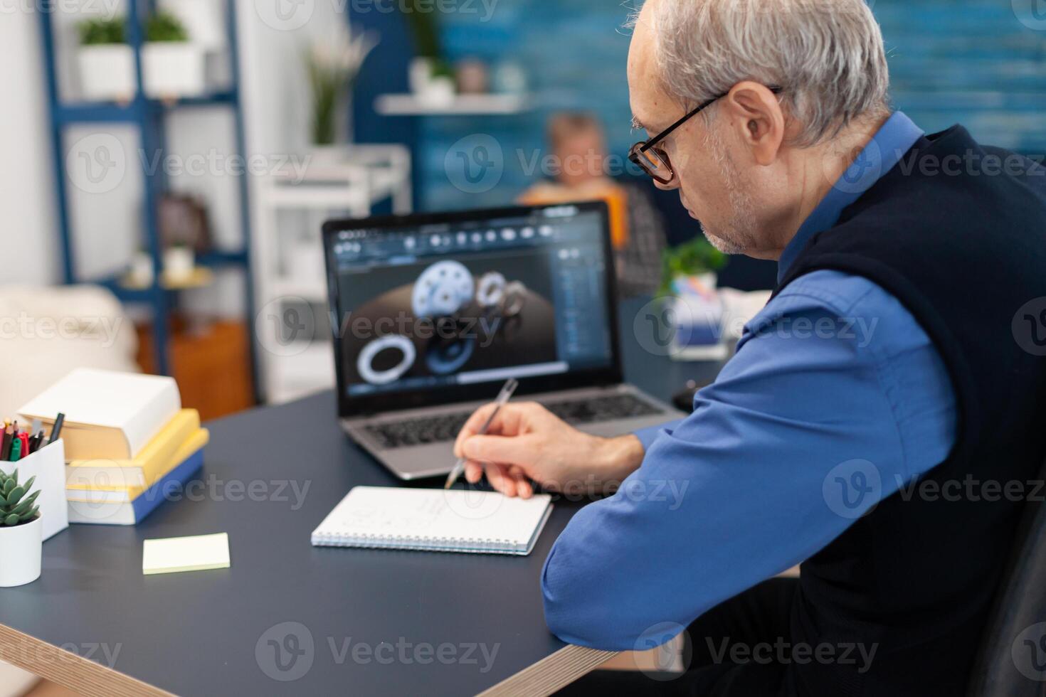 Smart thoughtful technician writing on notebook. Experienced engineer working from home office on mechanical project using laptop computer technology and wife is reading a book on sofa. photo