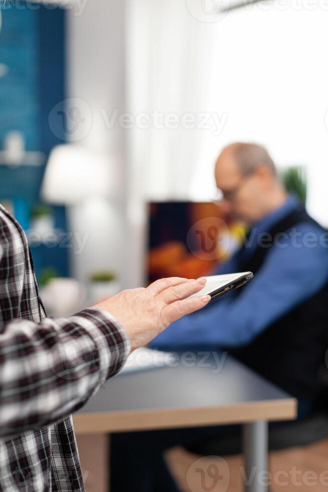 Close up of senior woman hand scrolling on digital device in home. Close up of elderly woman using moder technoloy tablet pc in home living room and husband working on laptop computer. photo