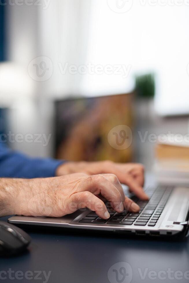 Close up of senior man hands typing on laptop keyboard. Elderly man entrepreneur in home workplace using portable computer sitting at desk while wife is holding tv remote photo