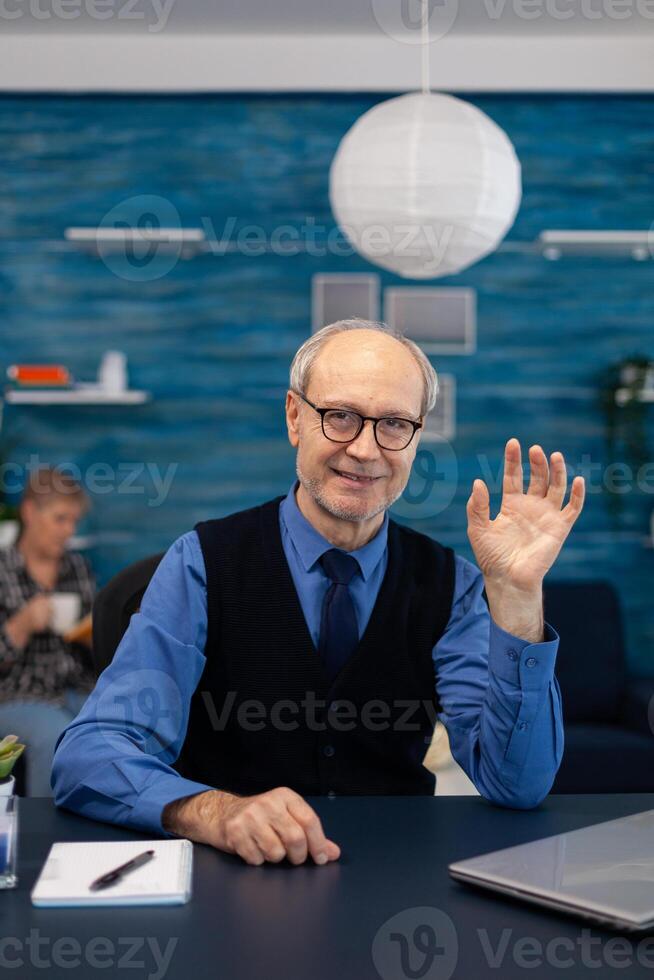 Smart senior businessman working on laptop wearing a tie and glasses. Elderly man entrepreneur in home workplace using portable computer sitting at desk while wife is holding tv remote. photo