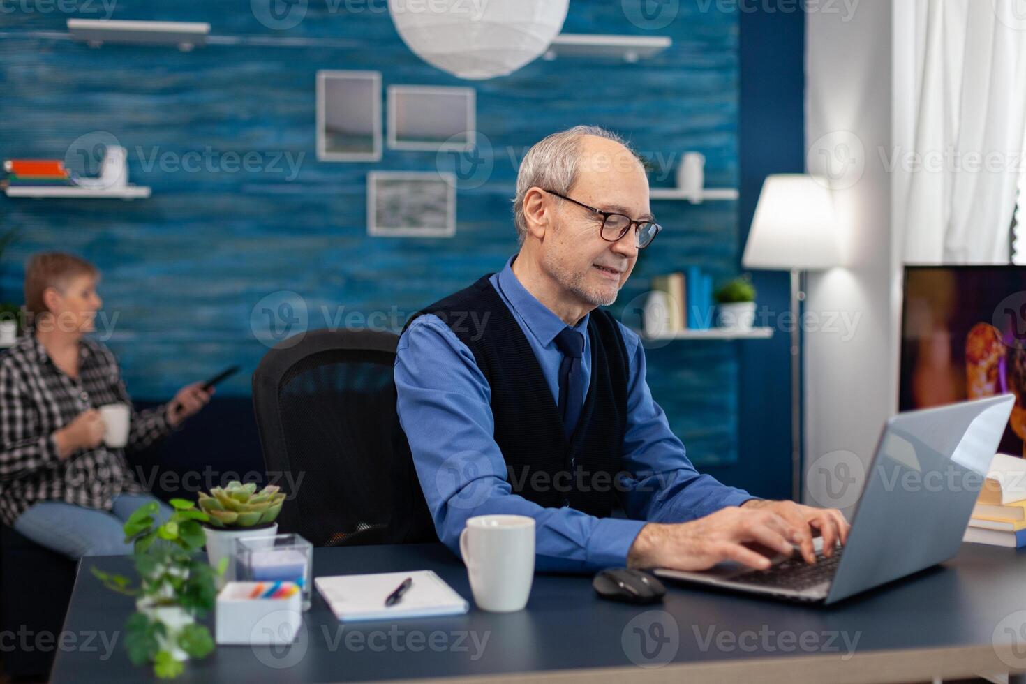 Portrait of senior man typing on laptop keyboard working on project. Elderly man entrepreneur in home workplace using portable computer sitting at desk while wife is holding tv remote. photo