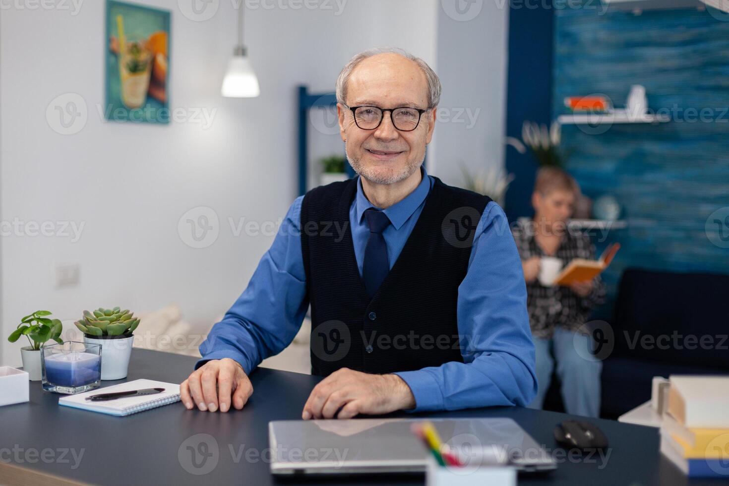 Cheerful senior businessman with gray hair. smiling at camera Elderly man entrepreneur in home workplace using portable computer sitting at desk while wife is reading a book sitting on sofa. photo