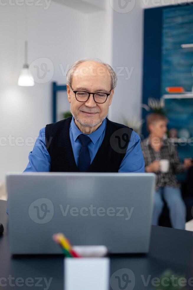 Old woman relaxing on couch holding a cup of coffee and husband is working. Elderly man entrepreneur in home workplace using portable computer sitting at desk while wife is holding tv remote. photo
