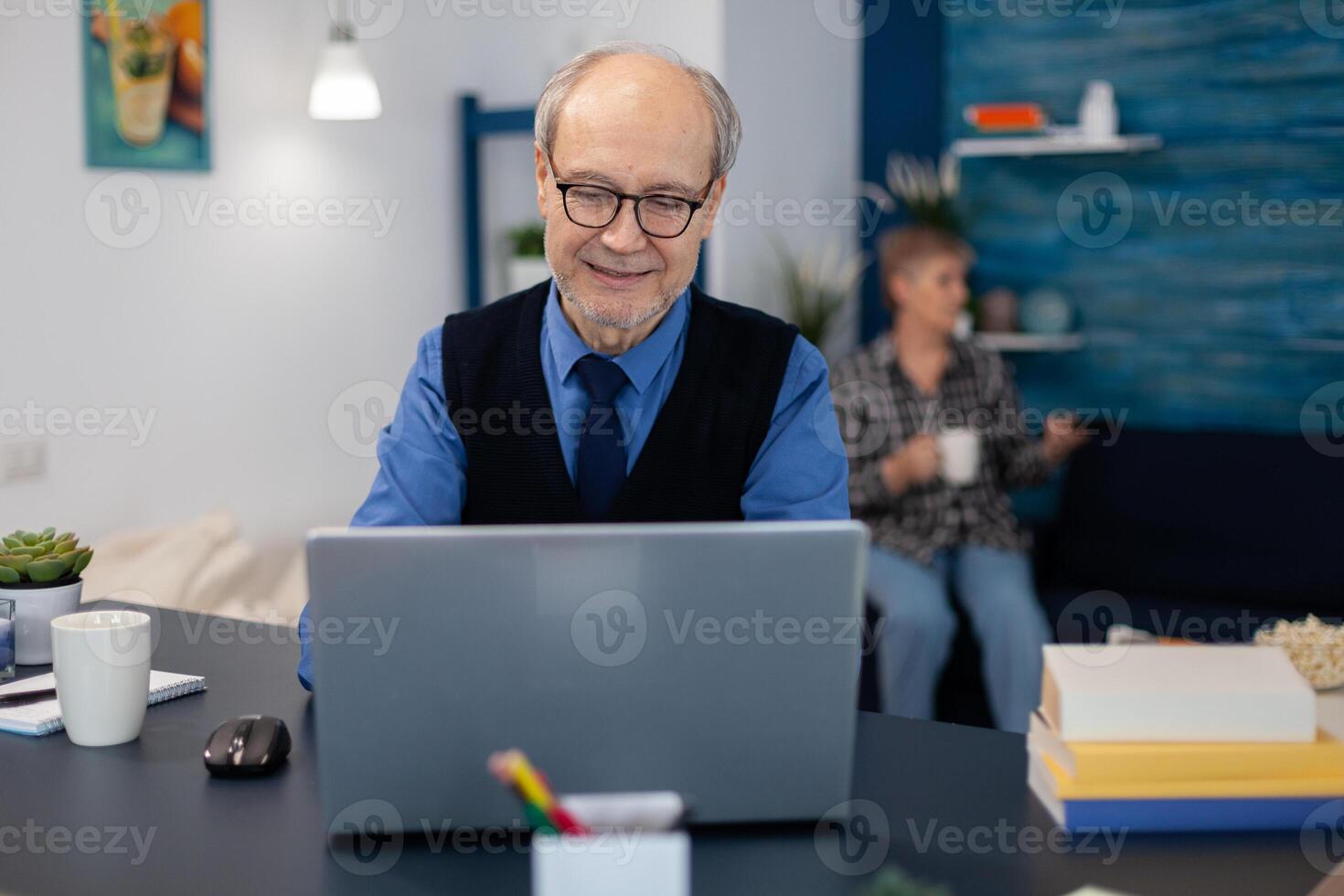Old woman relaxing on couch holding a cup of coffee and husband is working. Elderly man entrepreneur in home workplace using portable computer sitting at desk while wife is holding tv remote. photo
