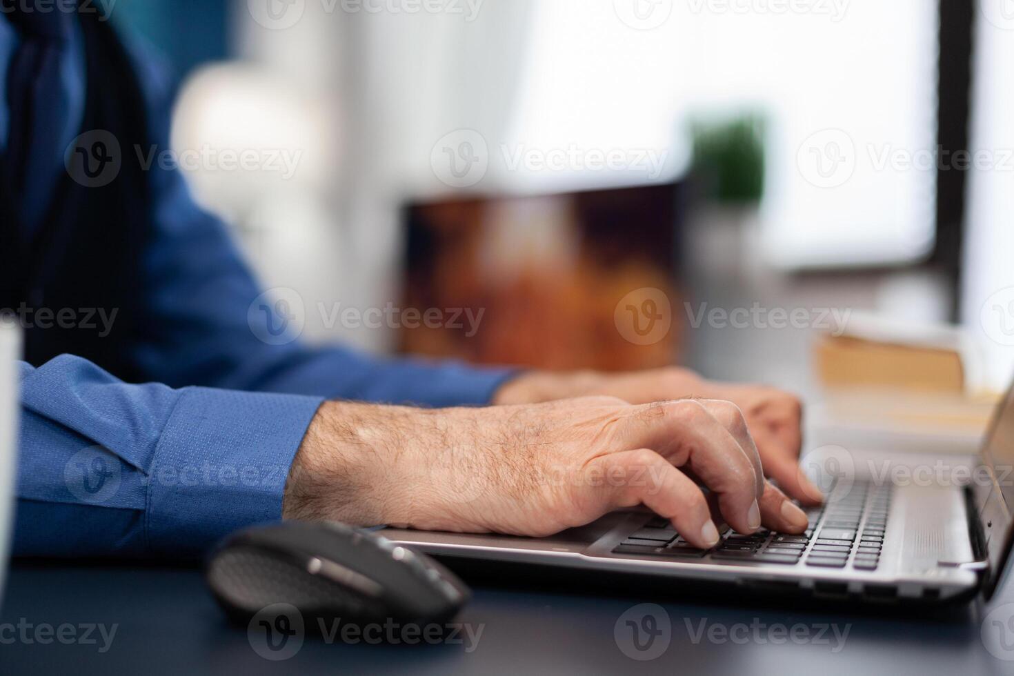 Close up of senior man hands typing on laptop keyboard. Elderly man entrepreneur in home workplace using portable computer sitting at desk while wife is holding tv remote photo
